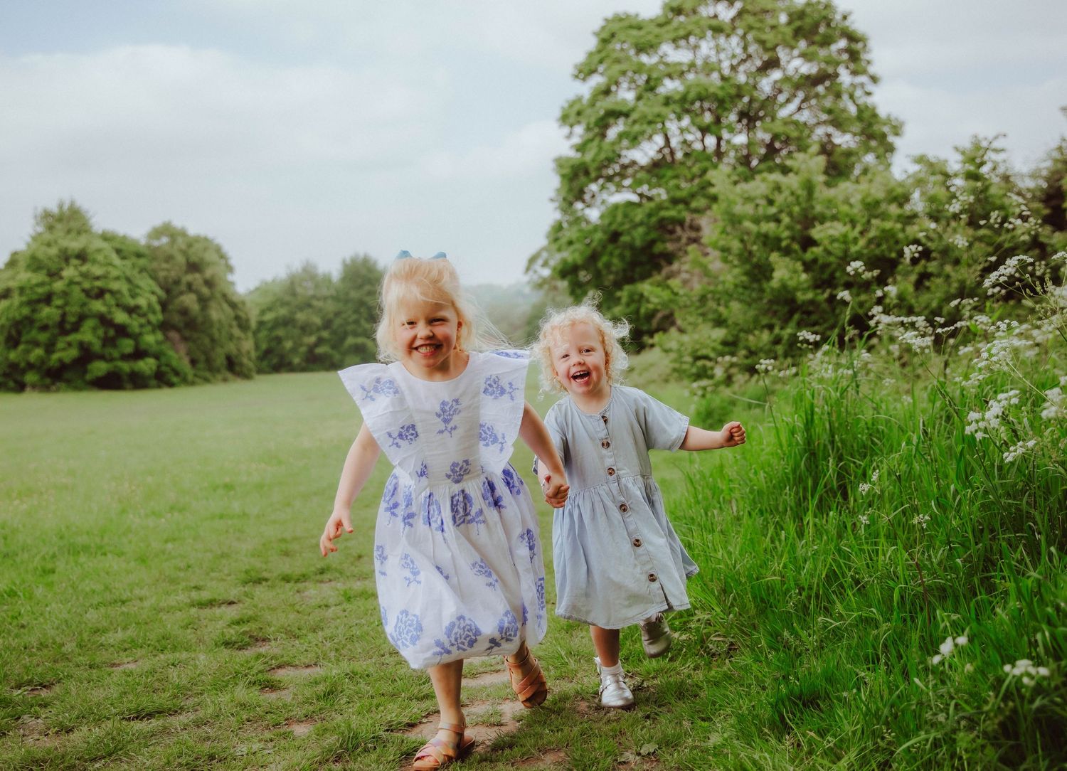 Two young girls in floral dresses laughing and running through a green meadow During a family photoshoot in South East London.