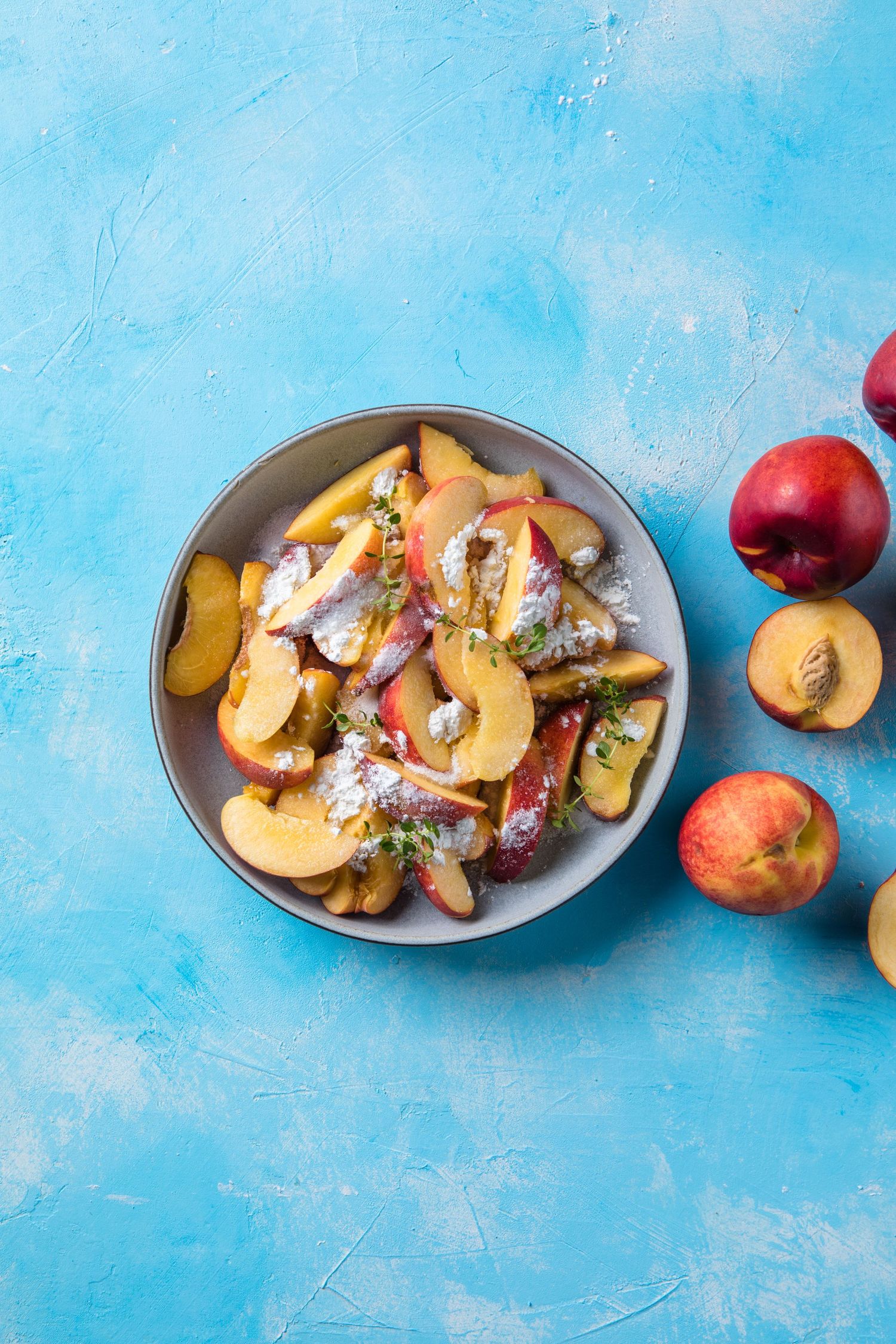 Fresh sliced nectarines in a bowl with powdered sugar on a blue background, with whole nectarines beside it.
