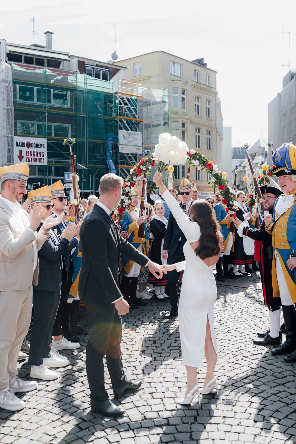 Bride raises bouquet as groom leads her through cheering crowd in traditional costumes on cobblestone street.