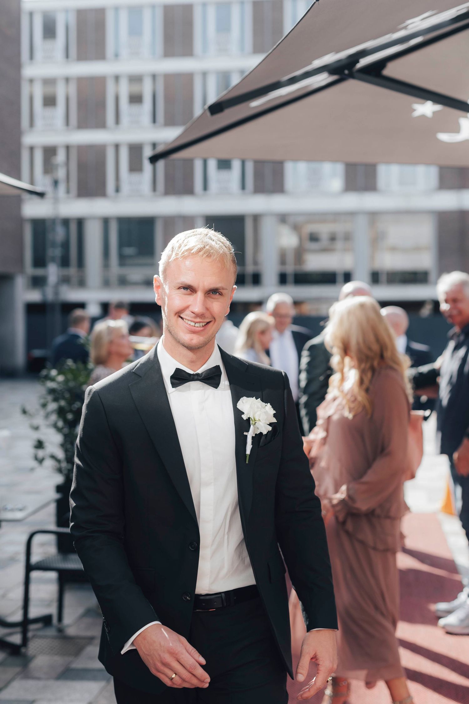 Smiling groom in black tuxedo with white boutonniere poses outdoors at wedding reception with guests in background.