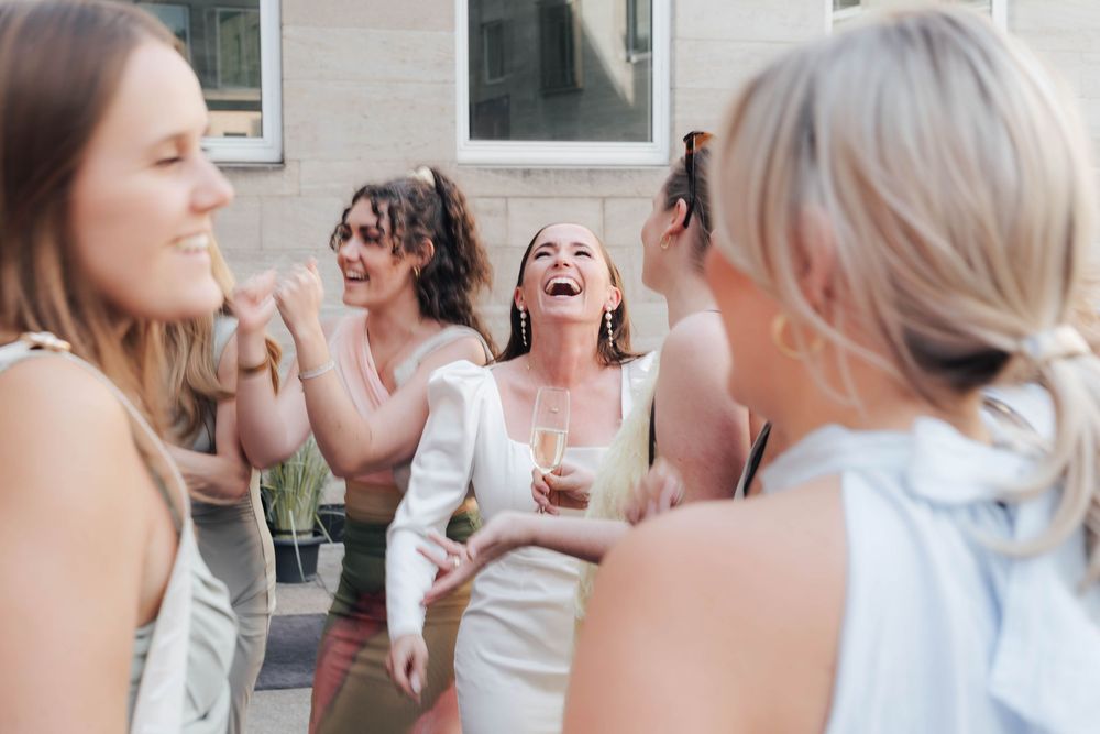Group of women laughing and celebrating outdoors, one in white holding champagne, likely a bridal party event.