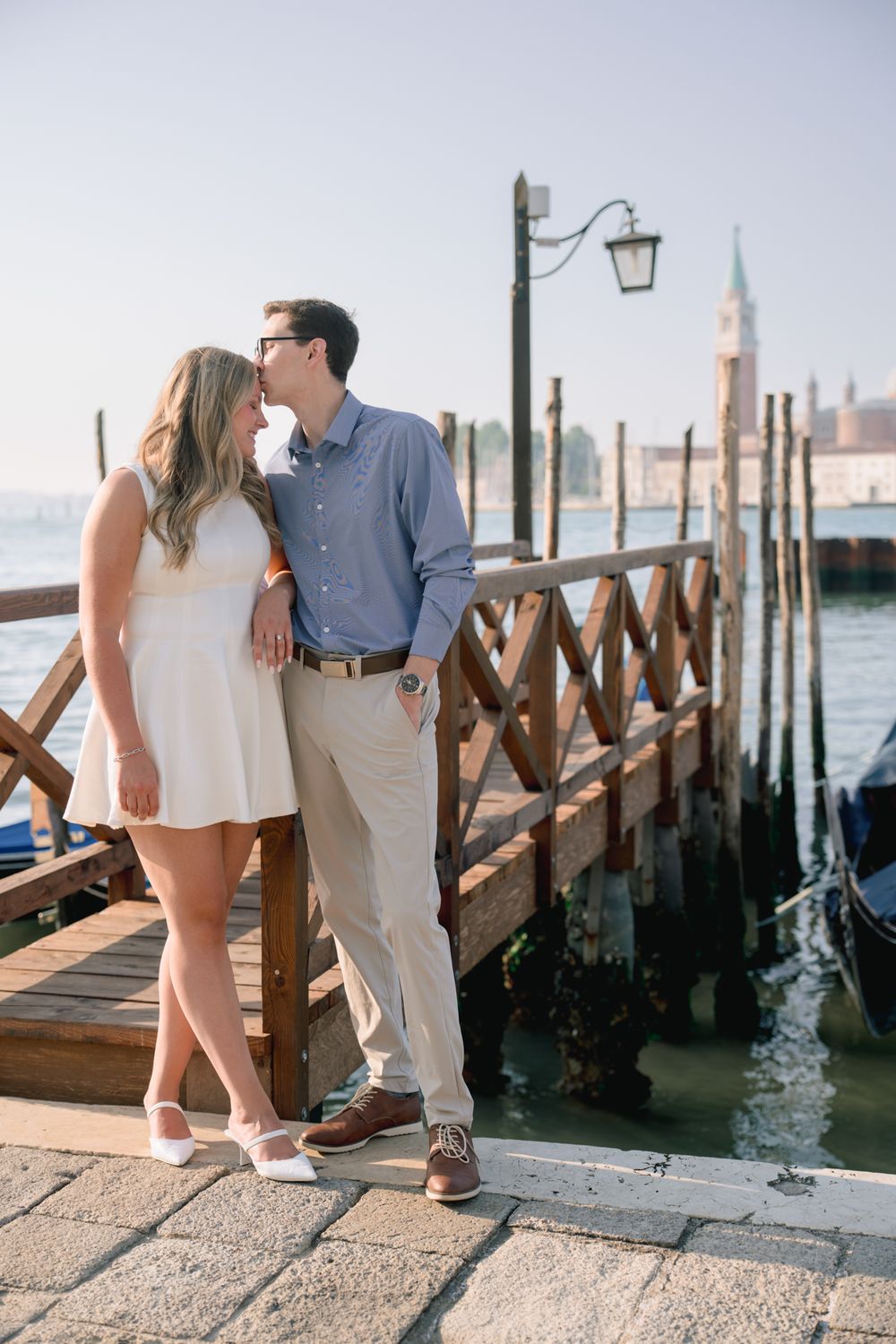 Couple sharing a romantic kiss on a wooden dock in Venice, Italy, with gondolas and historic architecture in the background.