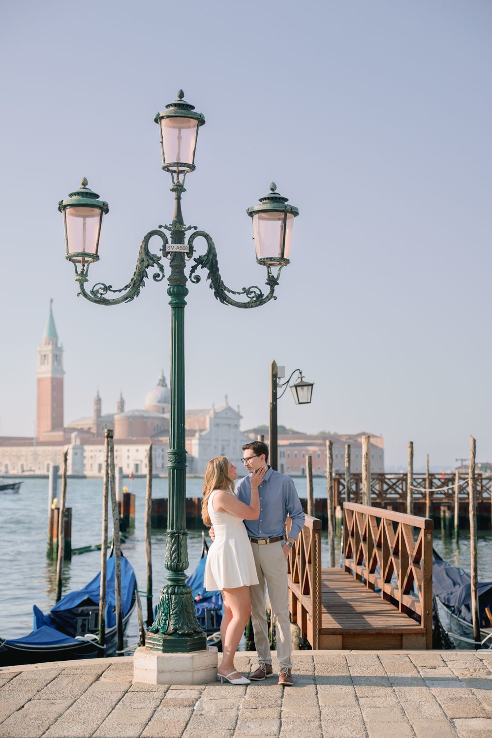 Couple embracing by ornate green lamppost on Venice waterfront with gondolas and San Giorgio Maggiore in background.