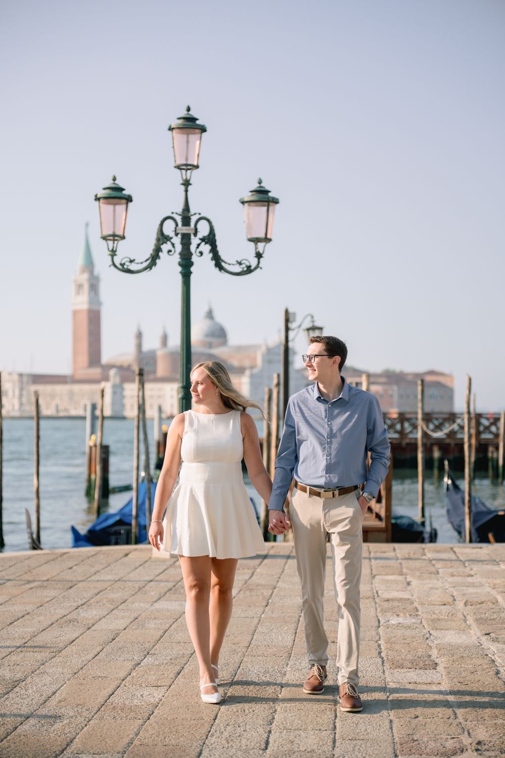 Couple holding hands walking along Venice waterfront promenade with gondolas and San Giorgio Maggiore in background.