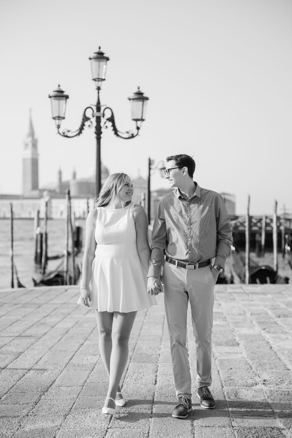 Black and white photo of a couple holding hands and walking along a Venice waterfront with gondolas and a lamp post.
