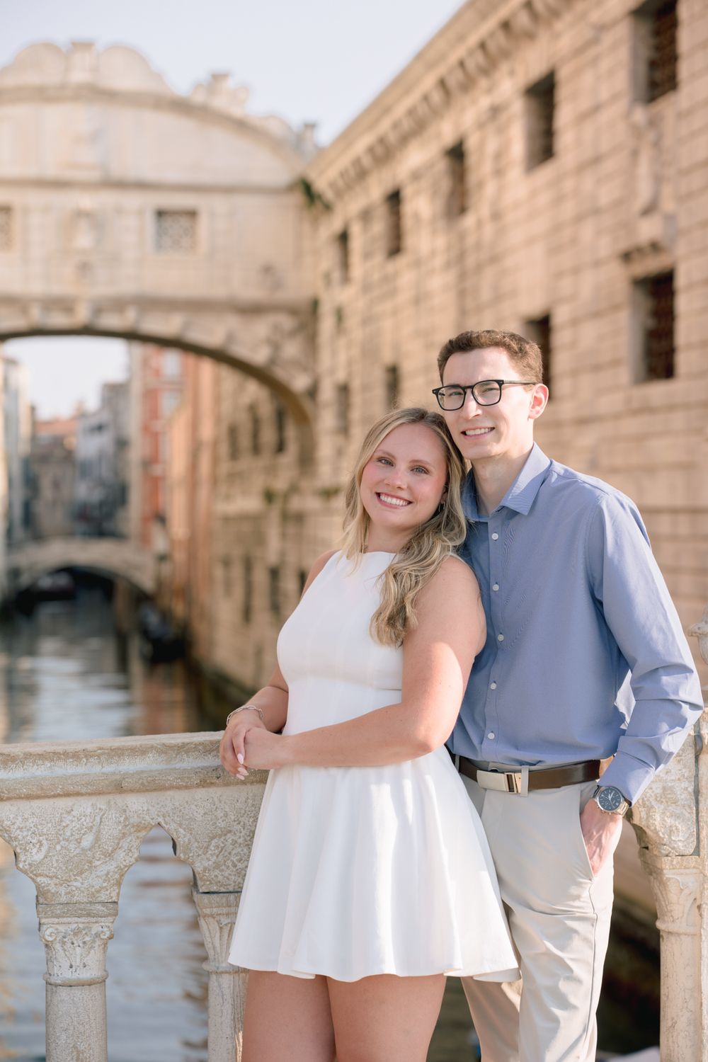 A smiling couple poses on a bridge over a Venice canal with the Bridge of Sighs in the background.