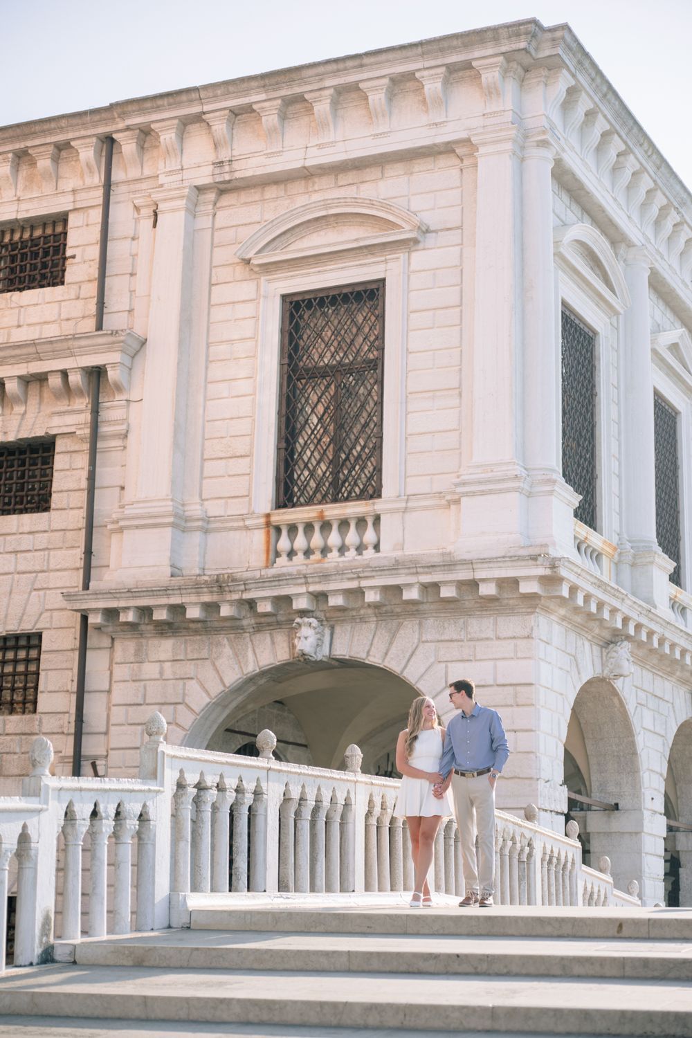 Couple standing on white marble steps of an elegant classical European building with arches and balustrades.