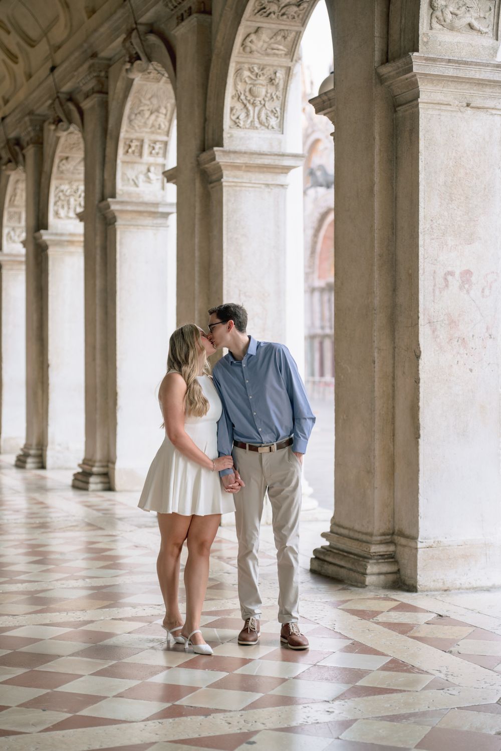 Couple sharing a romantic kiss under ornate marble arches on a checkered floor in Venice, Italy.