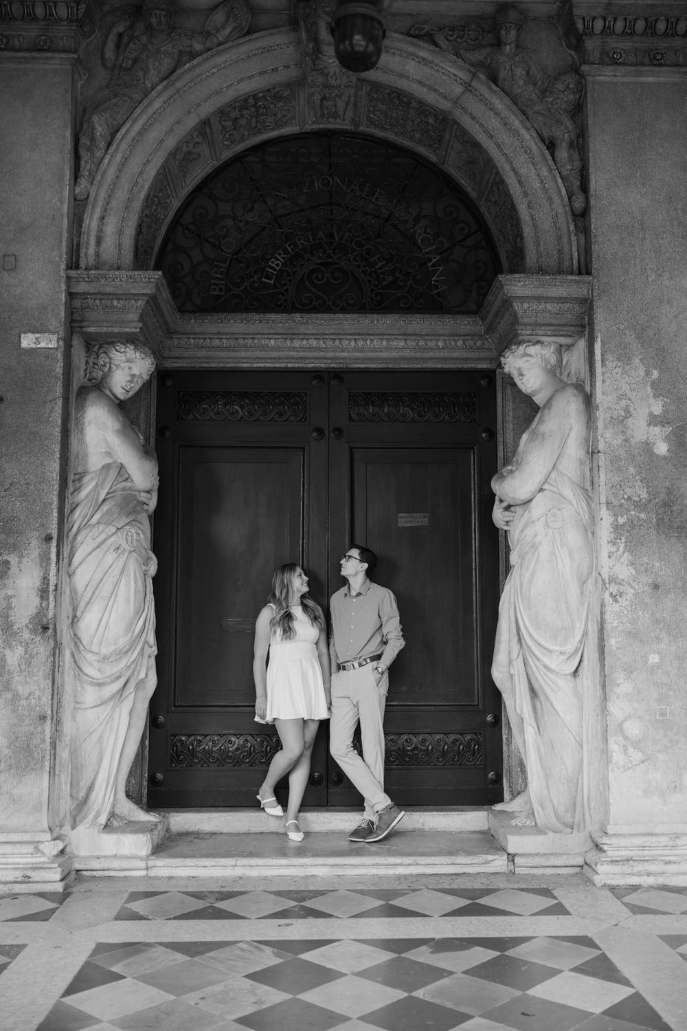 Black and white photo of a couple standing between classical statues at an ornate European doorway.
