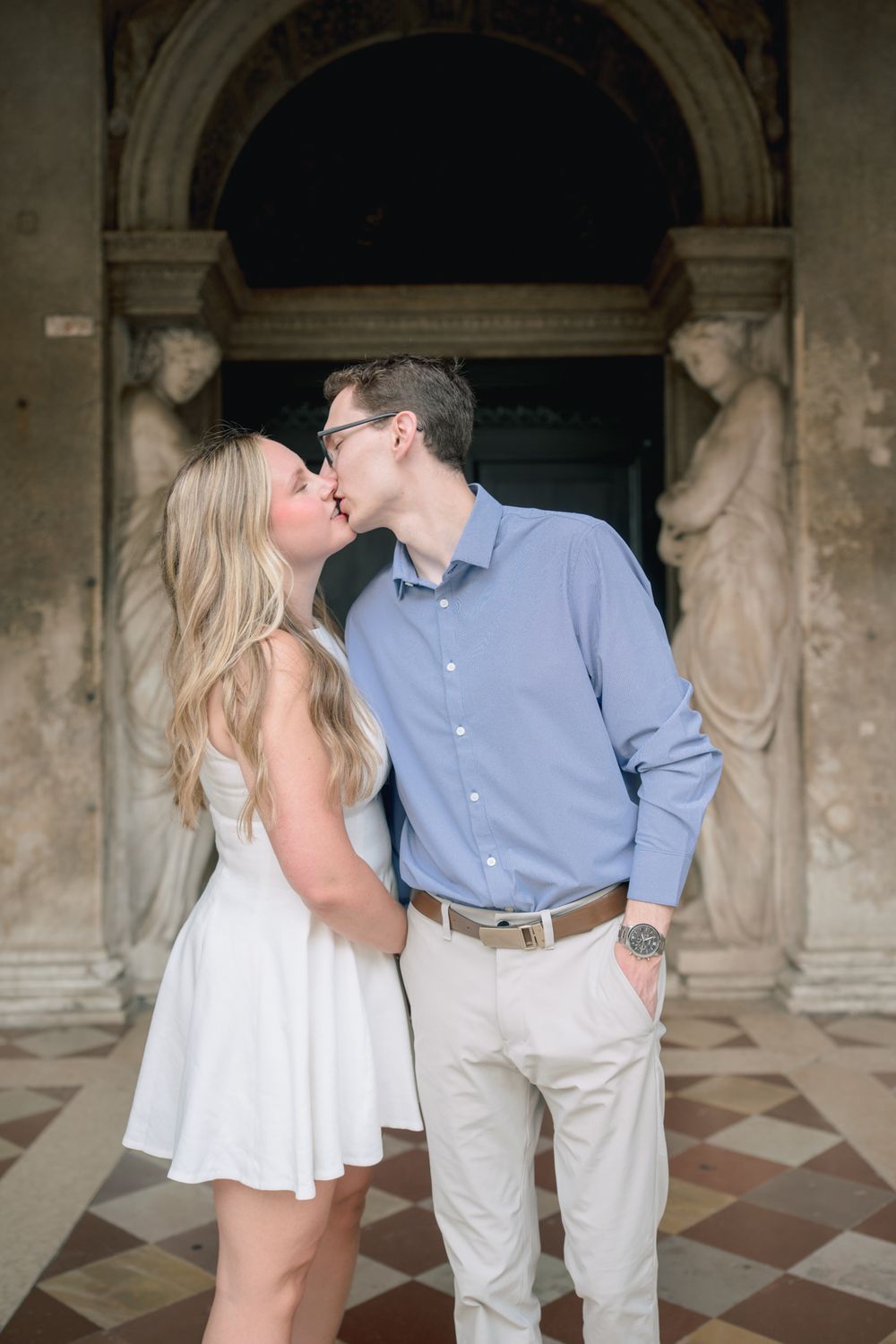 Couple sharing a romantic kiss in white dress and blue shirt before ornate stone archway with classical sculptures.