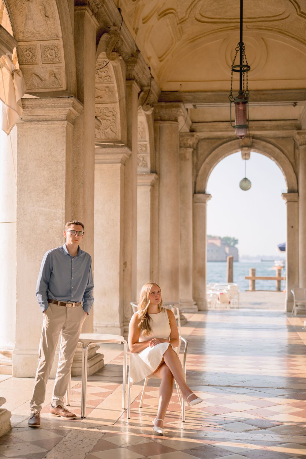 Couple posing in a sunlit Venetian arcade with marble columns and sea views.