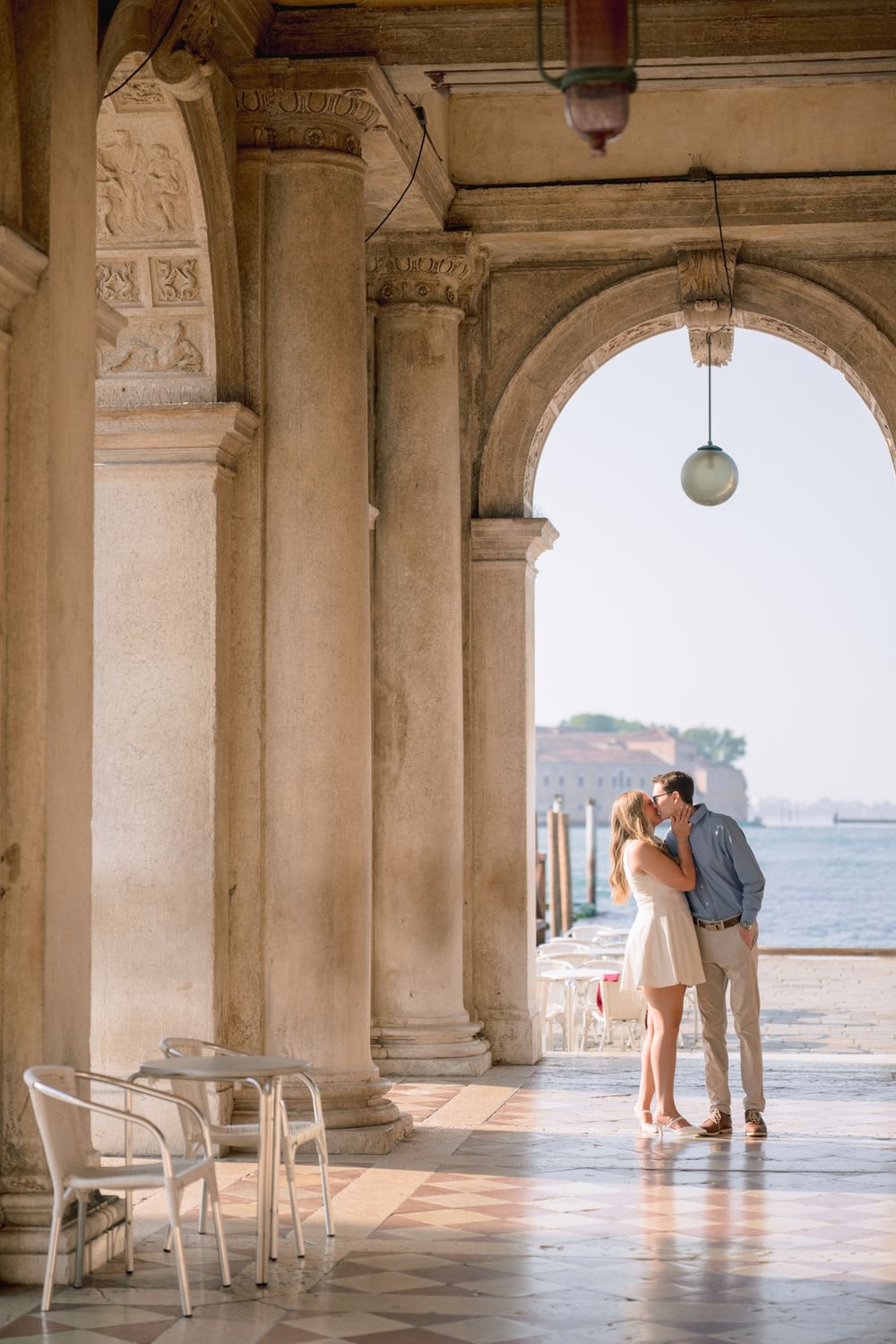 Couple kissing under elegant arches in Venice with waterfront views behind them.