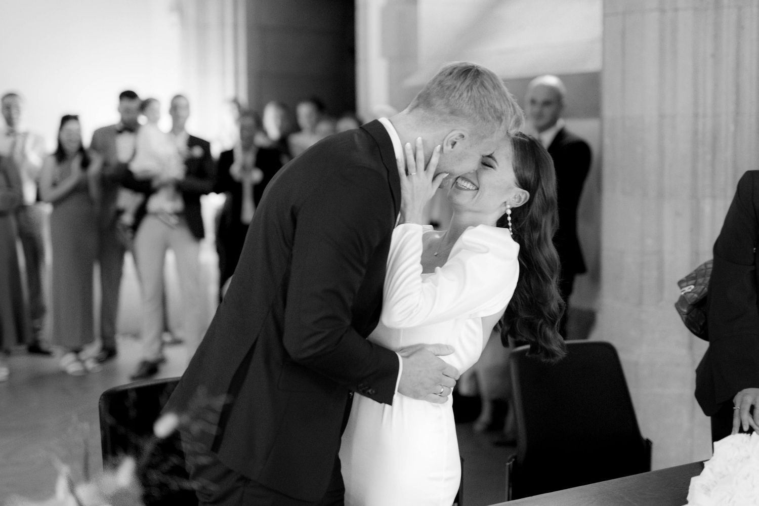 Black and white photo of a couple sharing their first kiss at a wedding ceremony with guests watching in the background.