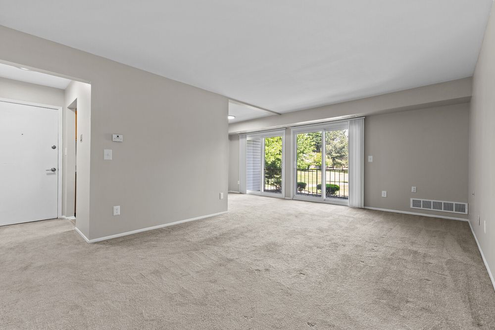 Open living room with beige carpet, neutral walls, sliding glass doors to a balcony, and a floor vent.