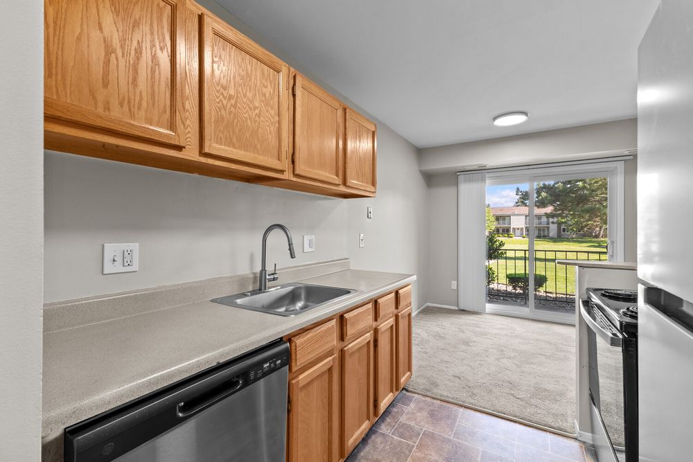 Bright apartment kitchen with oak cabinets, stainless steel dishwasher, sink, and sliding glass door leading to a balcony.