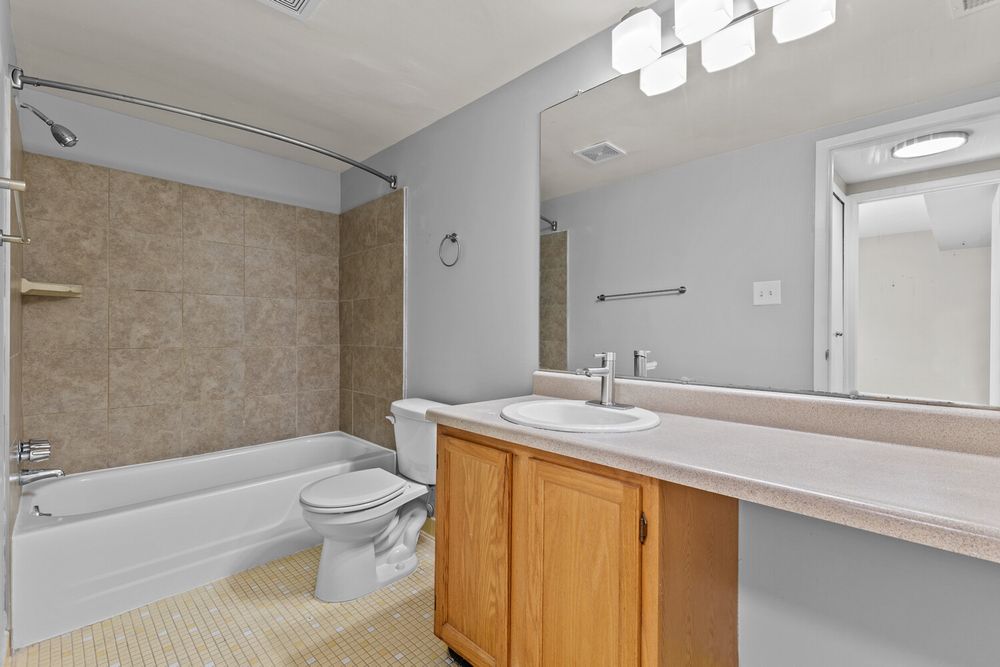 Full bathroom with oak vanity, large mirror, tiled shower-tub combo, and toilet on light tile floor.