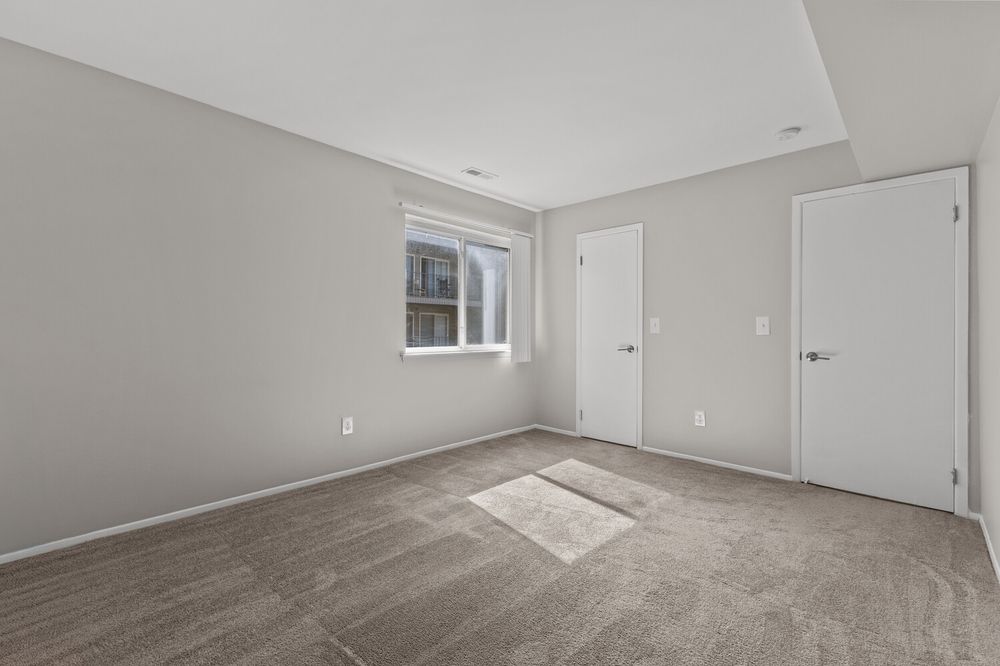 Empty bedroom with gray carpet, white walls, two white doors, and a window allowing natural light.