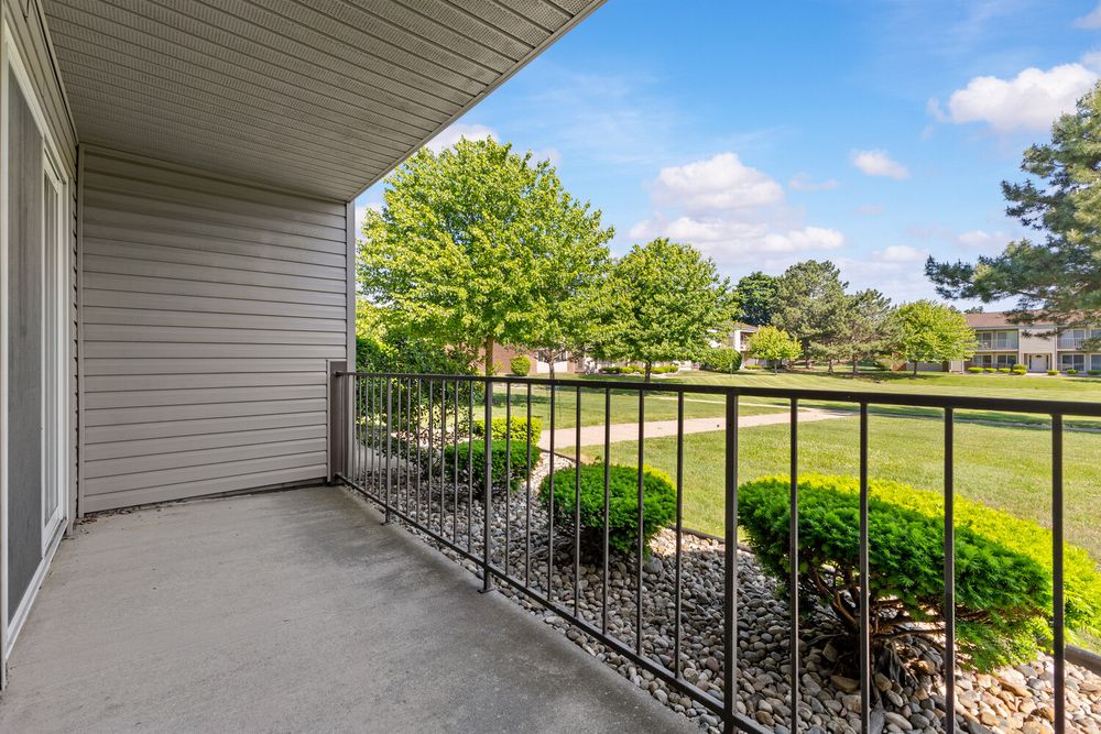 A private balcony with black iron railing overlooks a lush green lawn and mature trees under a blue sky.