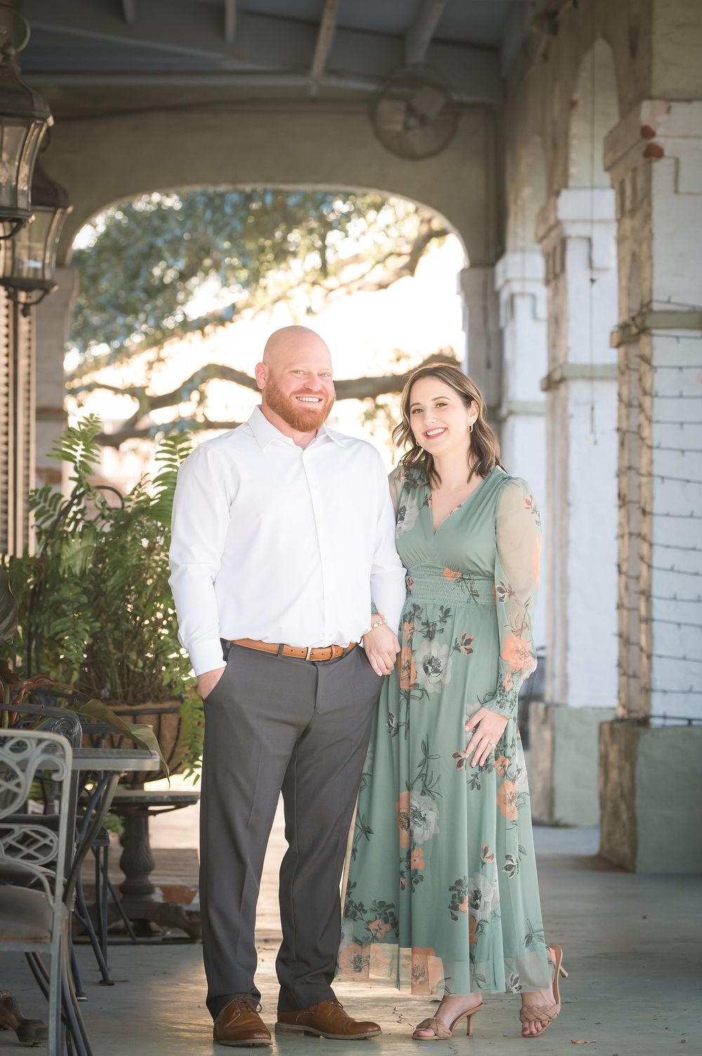 Smiling couple posing on a charming porch, man in white shirt and woman in green floral dress, surrounded by elegant architecture.