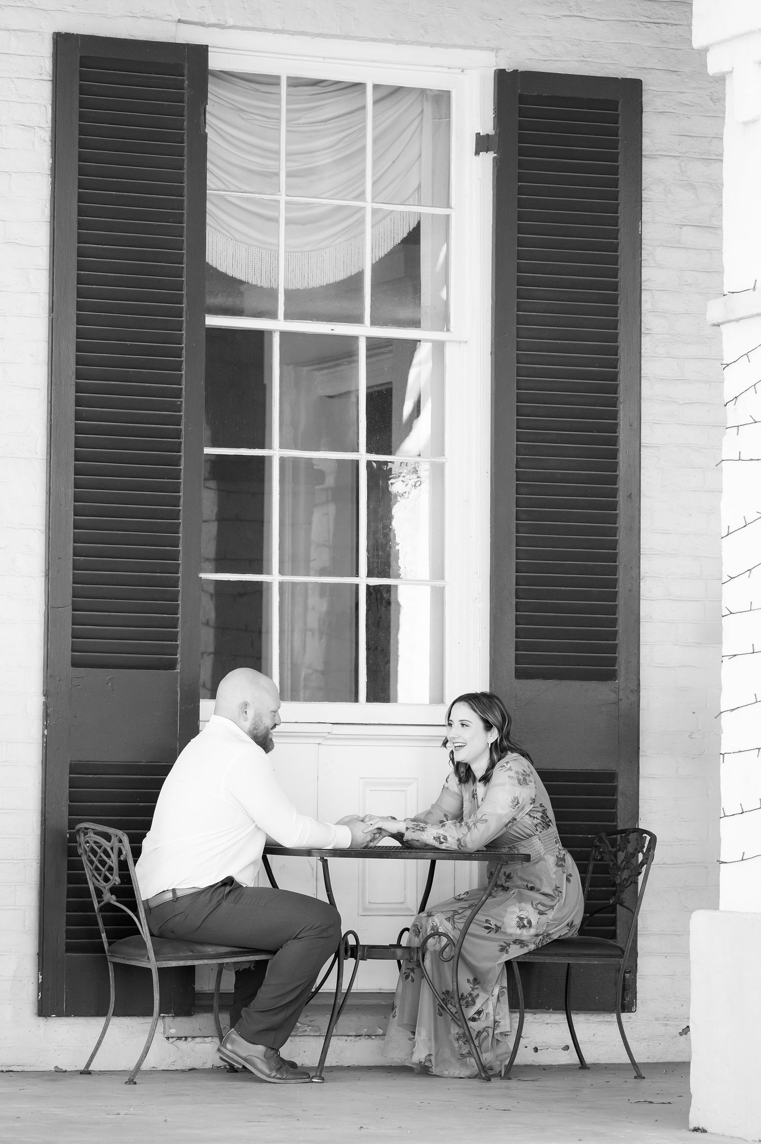 Black and white photo of a couple holding hands at a small iron table on a brick porch with shuttered windows.