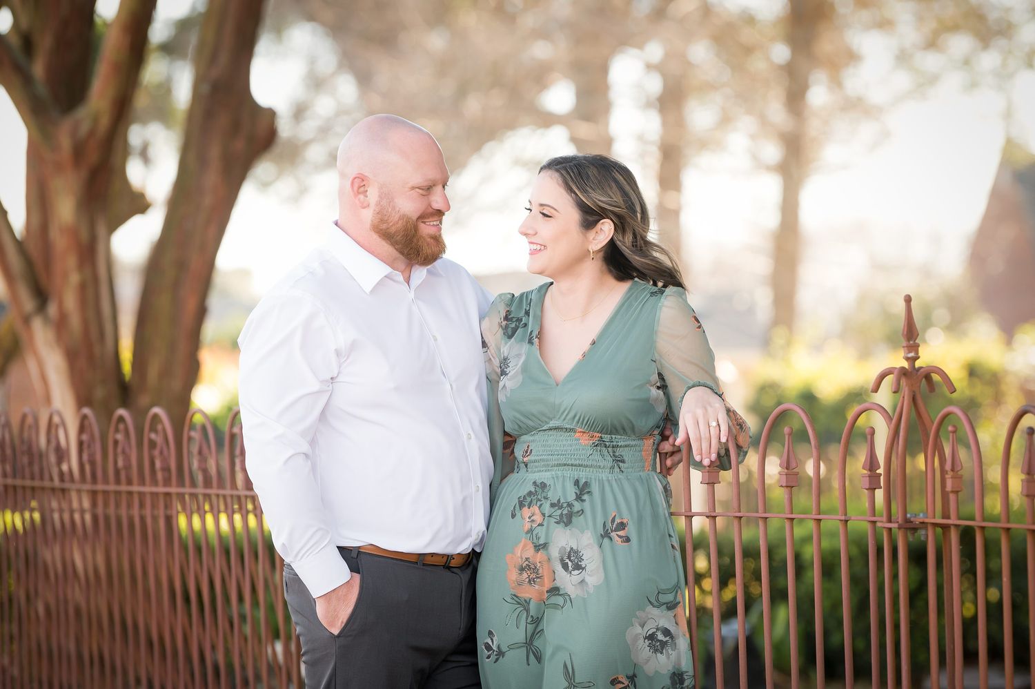 Couple smiling at each other outdoors near ornate iron fence, woman in floral green dress, man in white shirt.