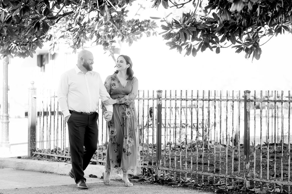 Couple walking and smiling together near iron fence under trees in black and white outdoor photo.