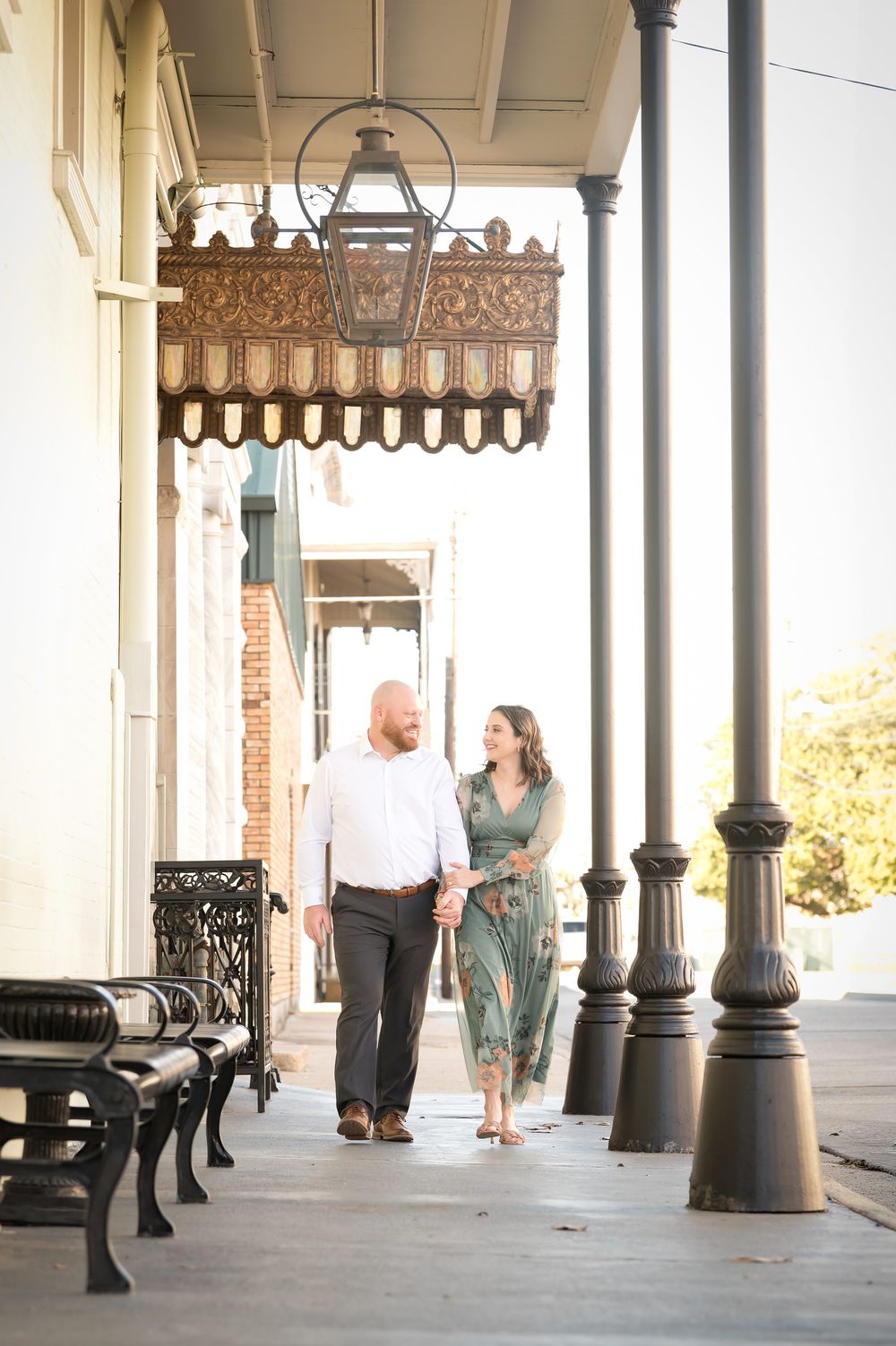 Couple holding hands walking along a historic Southern sidewalk with ornate lanterns and iron benches.
