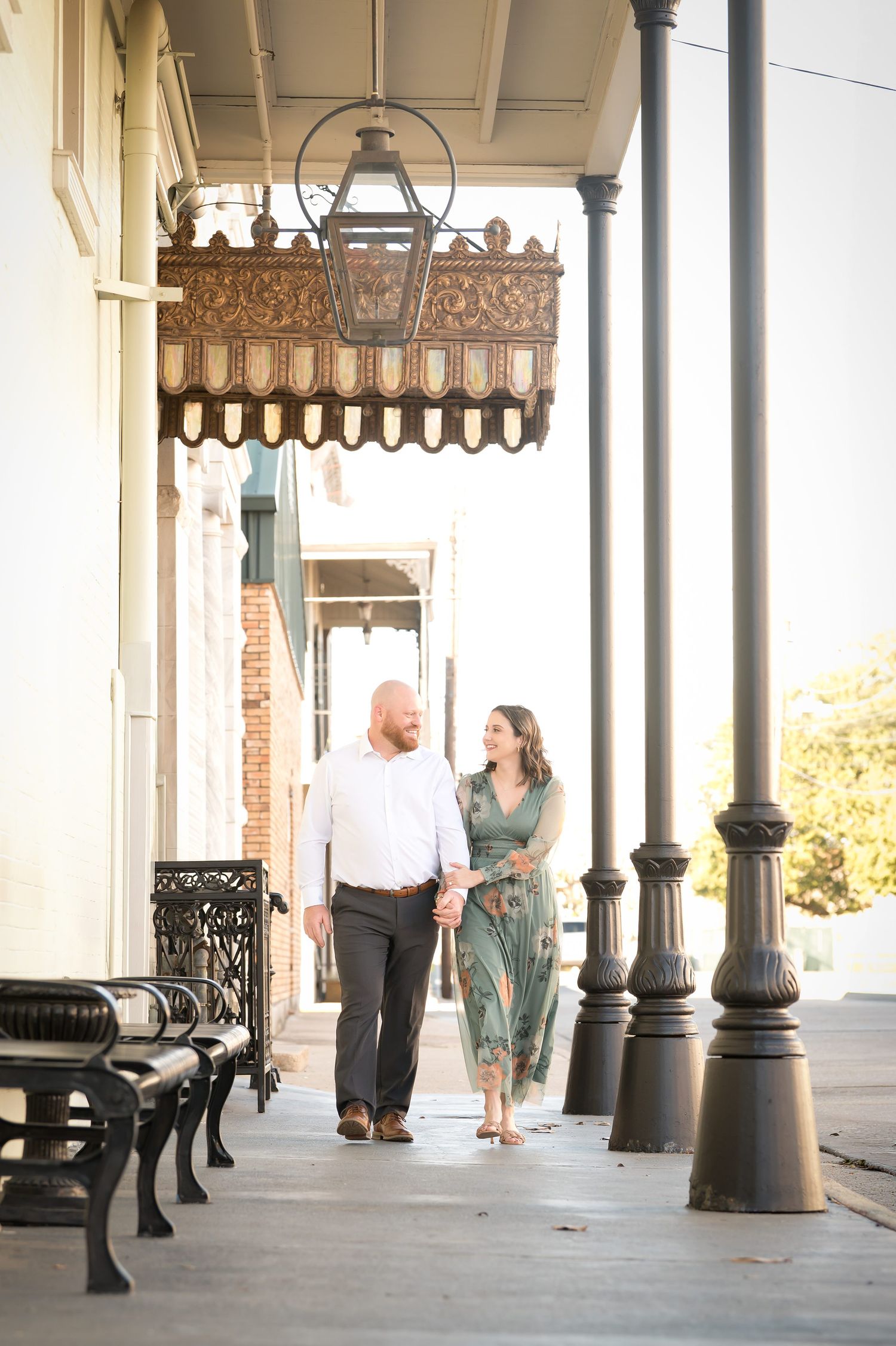Couple holding hands walking along a historic Southern sidewalk with ornate lanterns and iron benches.