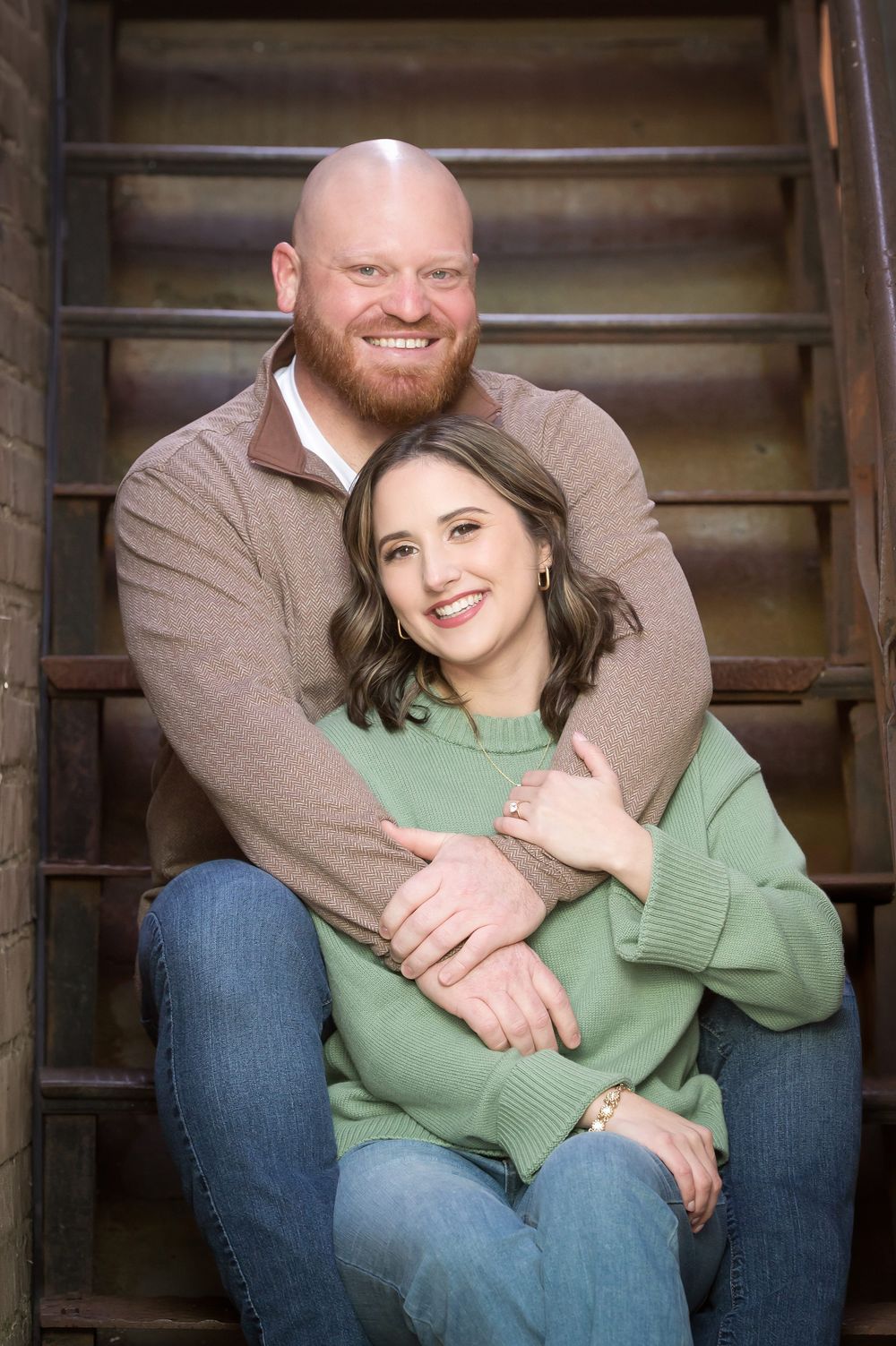 A smiling bald man with a red beard embraces a brunette woman in a green sweater on rustic wooden stairs.