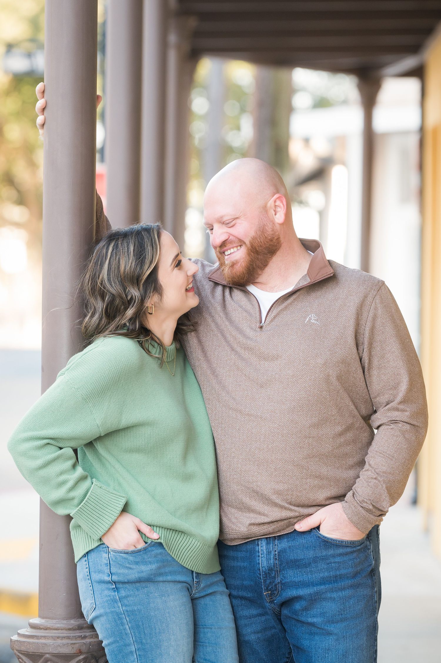 A smiling couple leans against a pole outdoors, woman in green sweater and man in brown pullover, both wearing jeans.