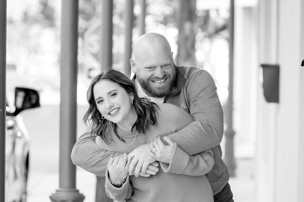 Black and white photo of a smiling couple embracing outdoors, man hugging woman from behind near pillars.