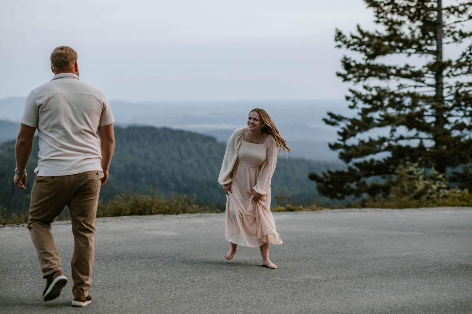 Couple walking on mountain overlook, woman in pink dress laughing, scenic forested valley in background.