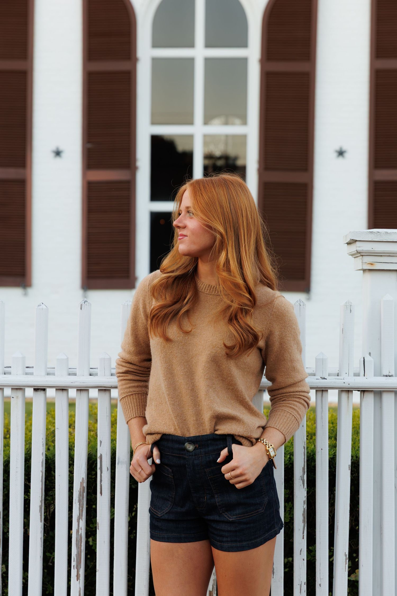 Red-haired woman in camel sweater and dark shorts standing by white picket fence outside historic building.