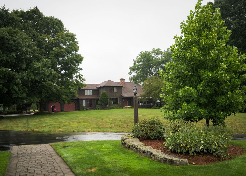 A large brick estate home surrounded by lush green trees and manicured lawn on a cloudy day.