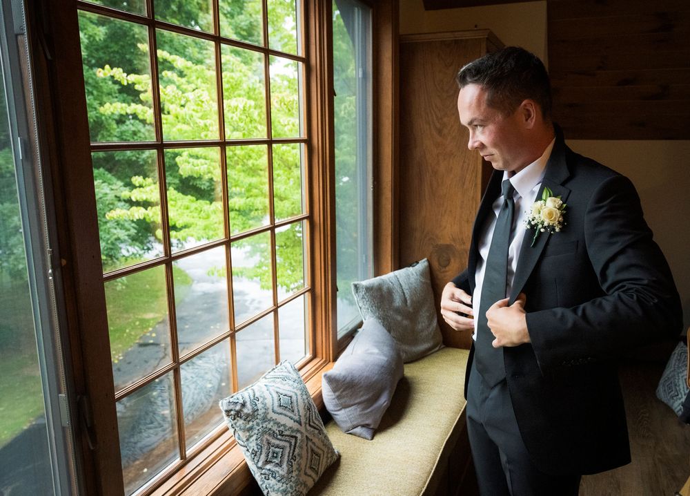 Groom in black suit adjusting boutonniere by rustic window with green trees visible outside on wedding day.