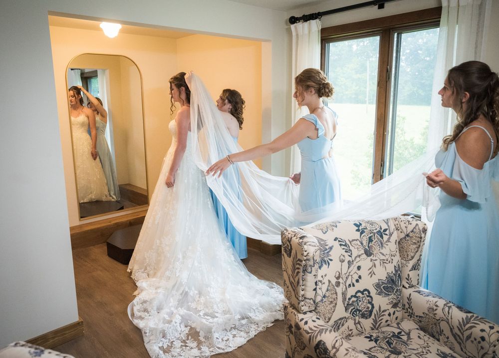Bridesmaids helping bride with lace wedding gown and veil in a rustic bridal suite before the ceremony.