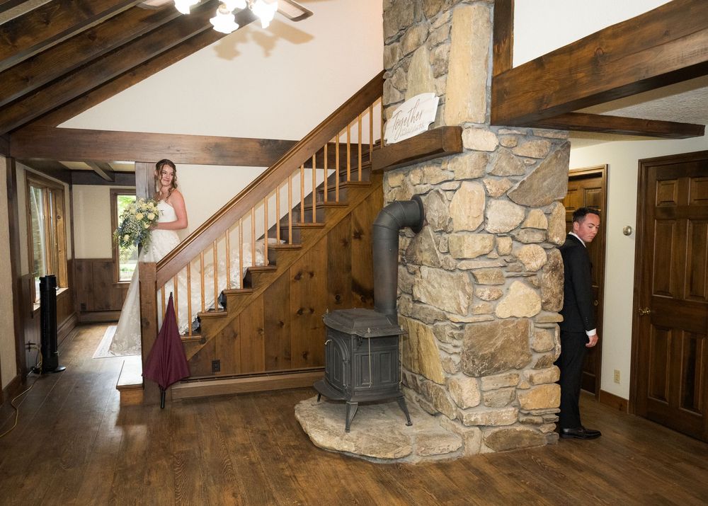 Bride descends wooden staircase past stone fireplace as groom waits below in rustic wedding venue.