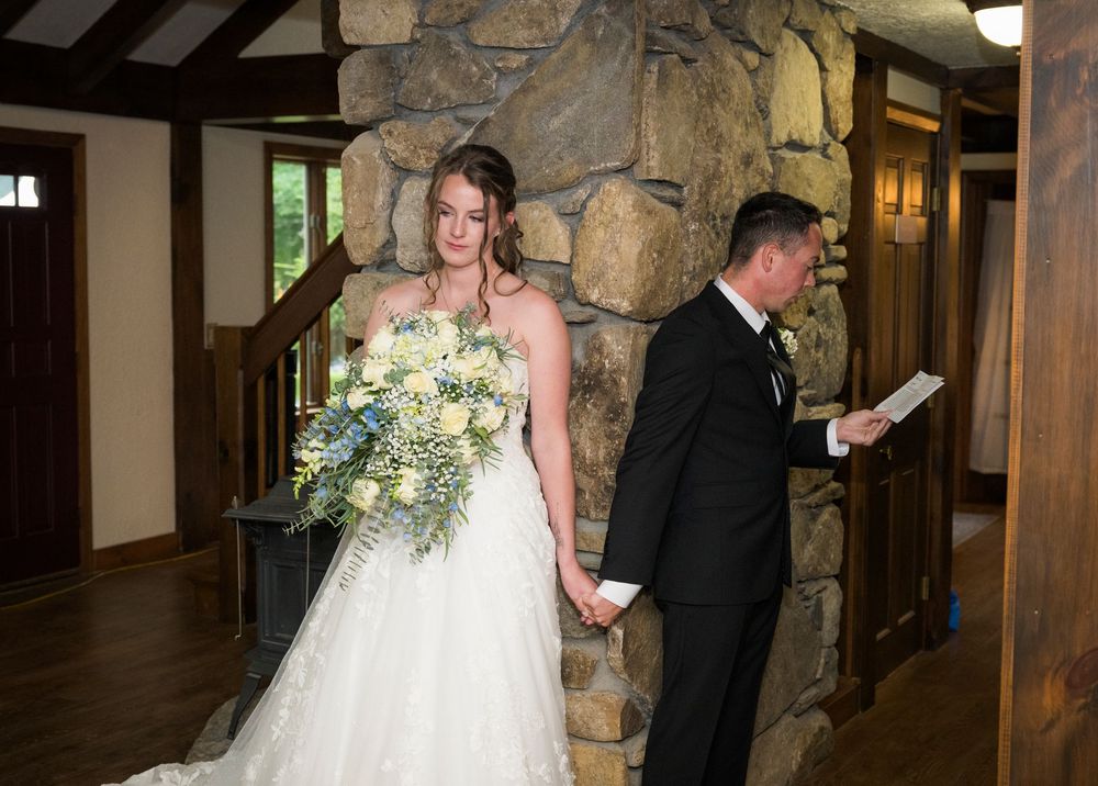 A bride holding a bouquet stands with a man in a tuxedo near a stone fireplace during a wedding ceremony.