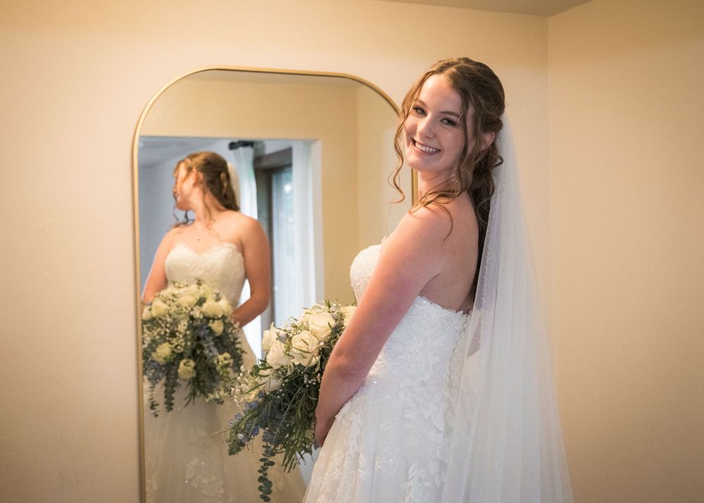 Smiling bride in white lace gown holding bouquet poses in front of mirror.