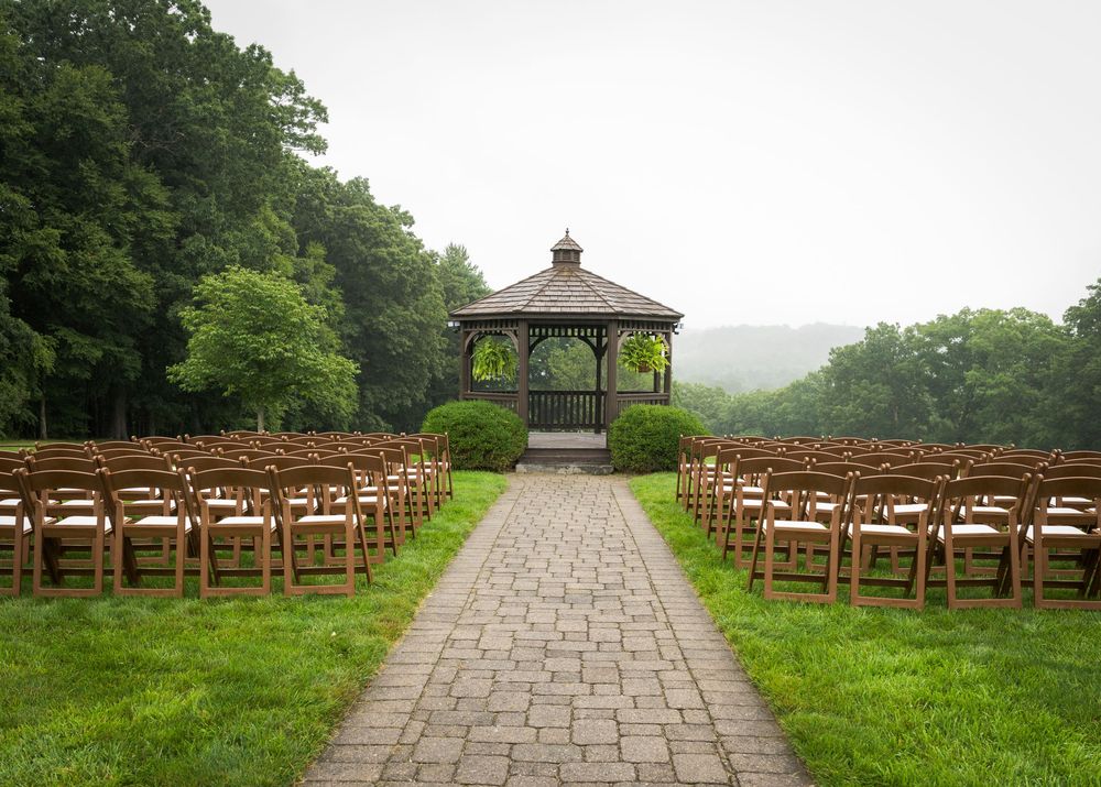Outdoor wedding ceremony setup with wooden chairs lining a brick pathway leading to a gazebo surrounded by lush greenery.