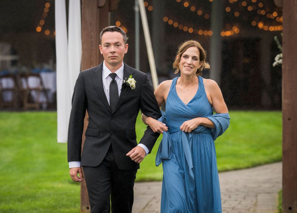 A groomsman in a black suit escorts a woman in a blue dress down the wedding aisle outdoors.