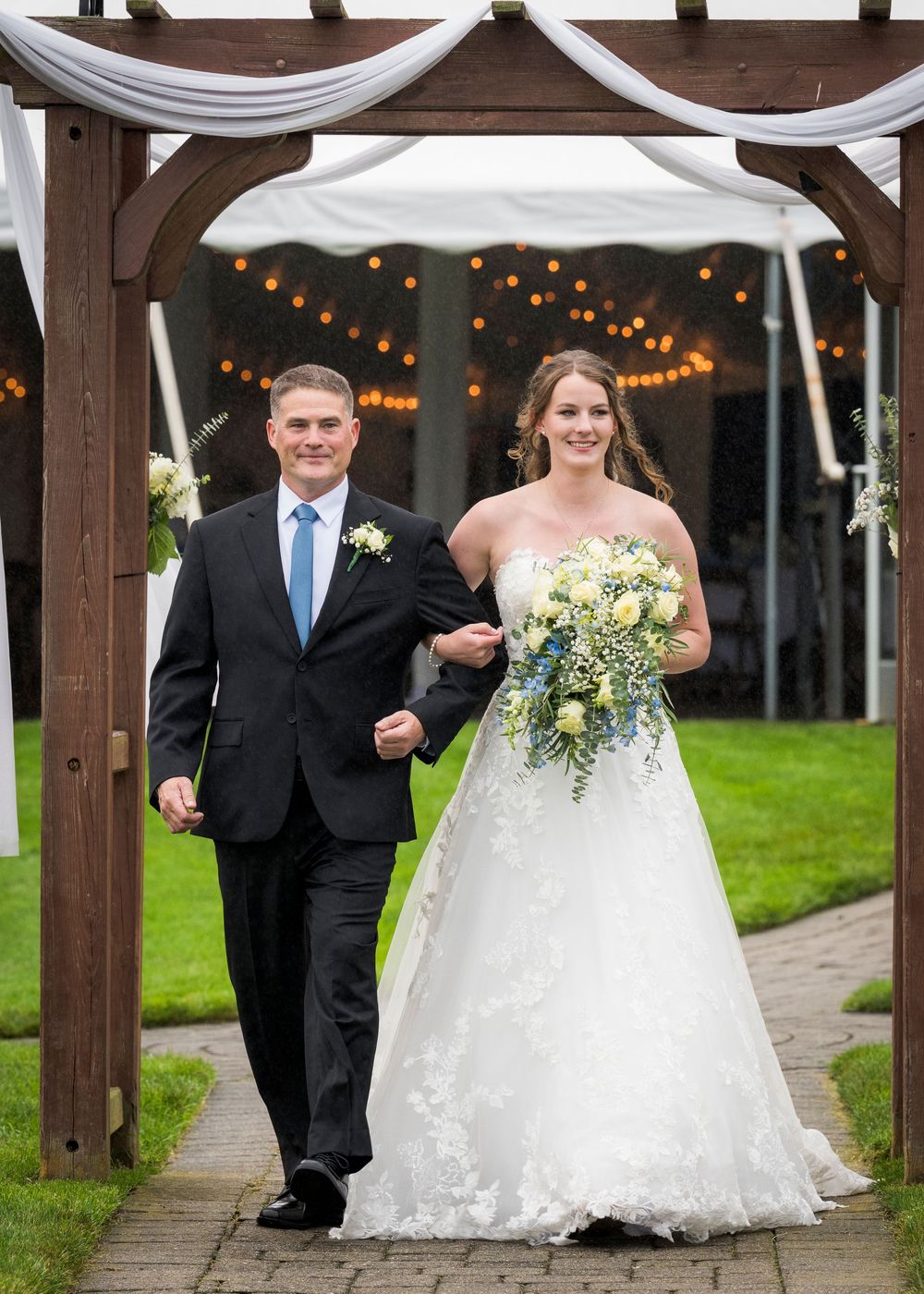 A groom in a black suit walks a bride in a white lace gown down the aisle under a wooden arbor.