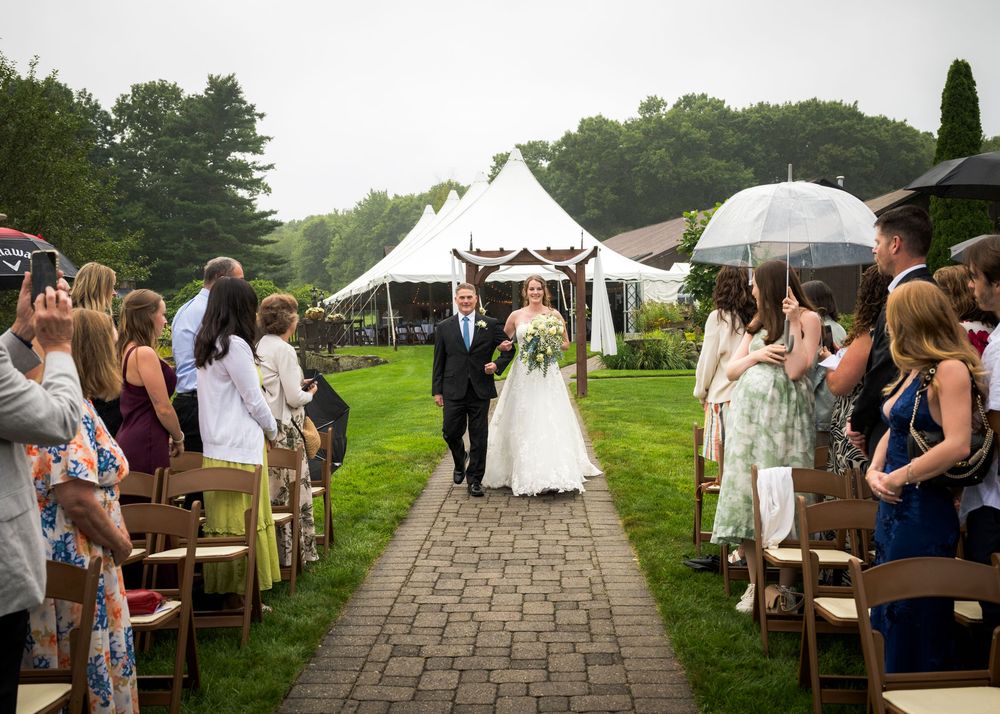 A bride walks down an outdoor aisle with guests holding umbrellas on a rainy wedding day.