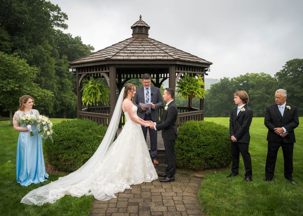 Bride and groom exchange vows at an outdoor gazebo wedding ceremony with bridal party on a lush green lawn.