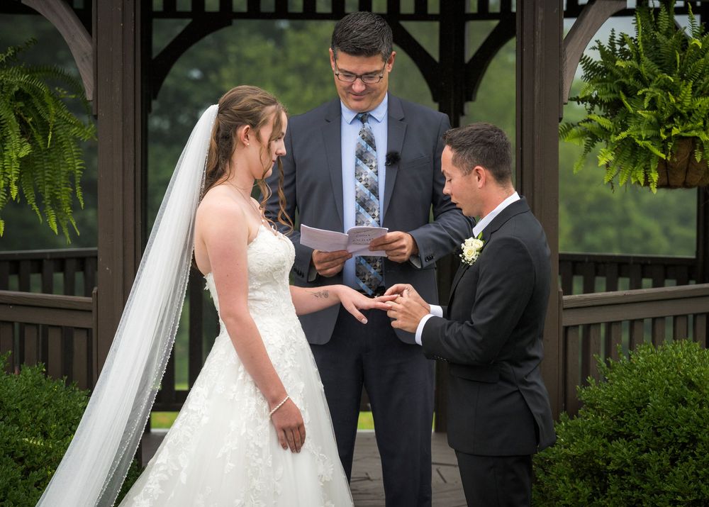A groom places a ring on the bride's finger during an outdoor wedding ceremony in a gazebo.