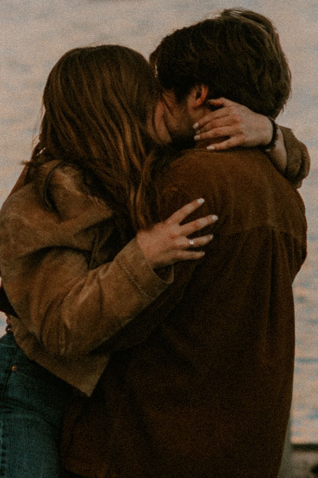 A couple shares a romantic embrace and kiss outdoors, wearing brown jackets in soft warm tones.