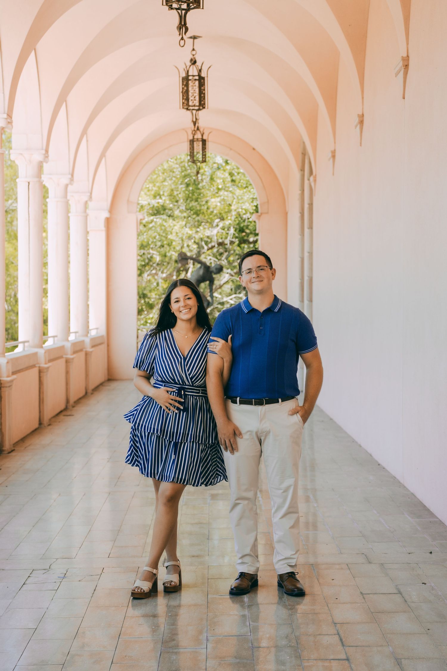 Pregnant couple posing under arched colonnade, woman in blue striped dress, man in blue polo shirt, pink architectural corridor.