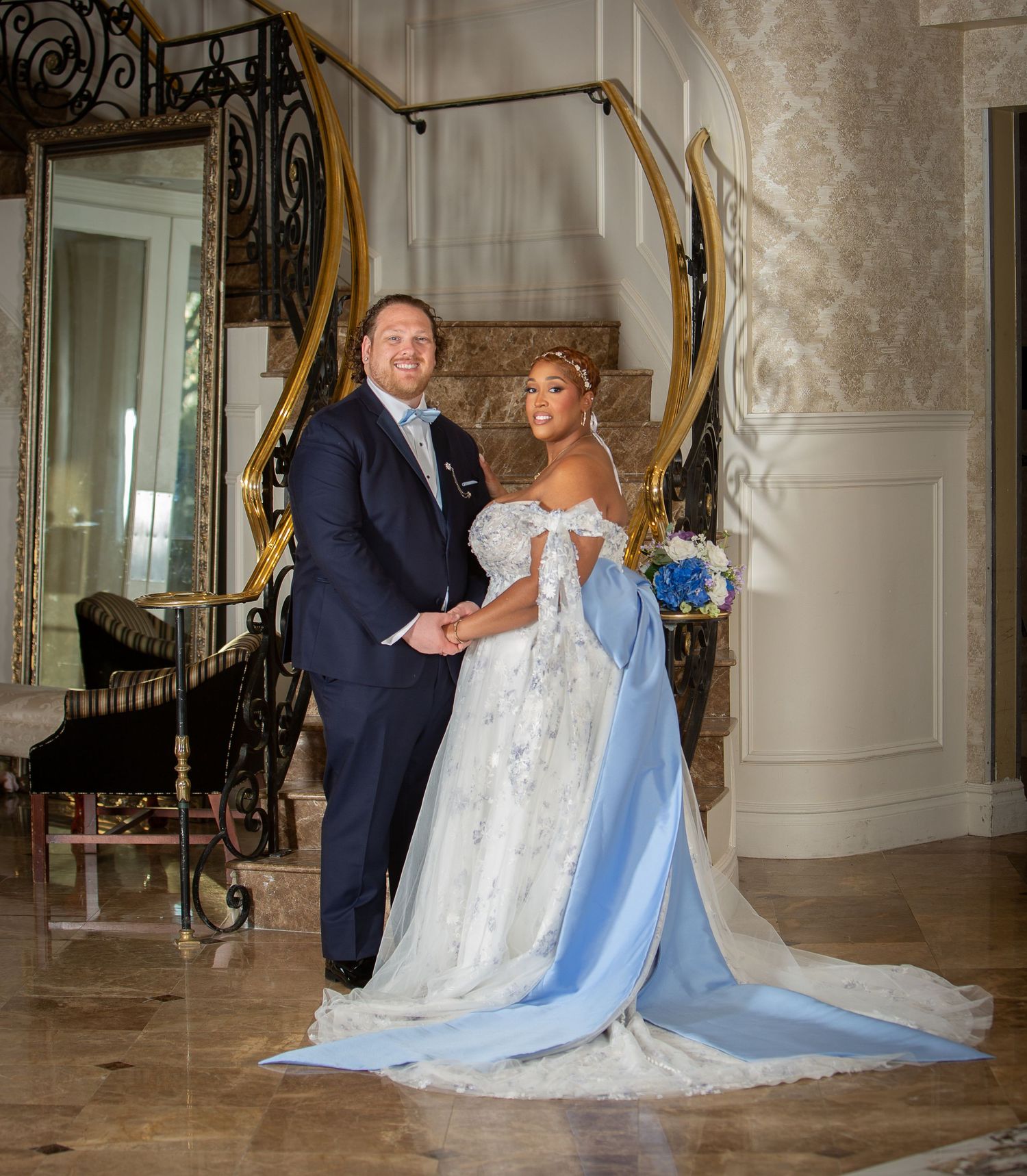 Bride in blue and white gown with groom in navy suit holding hands in elegant gold staircase venue.
