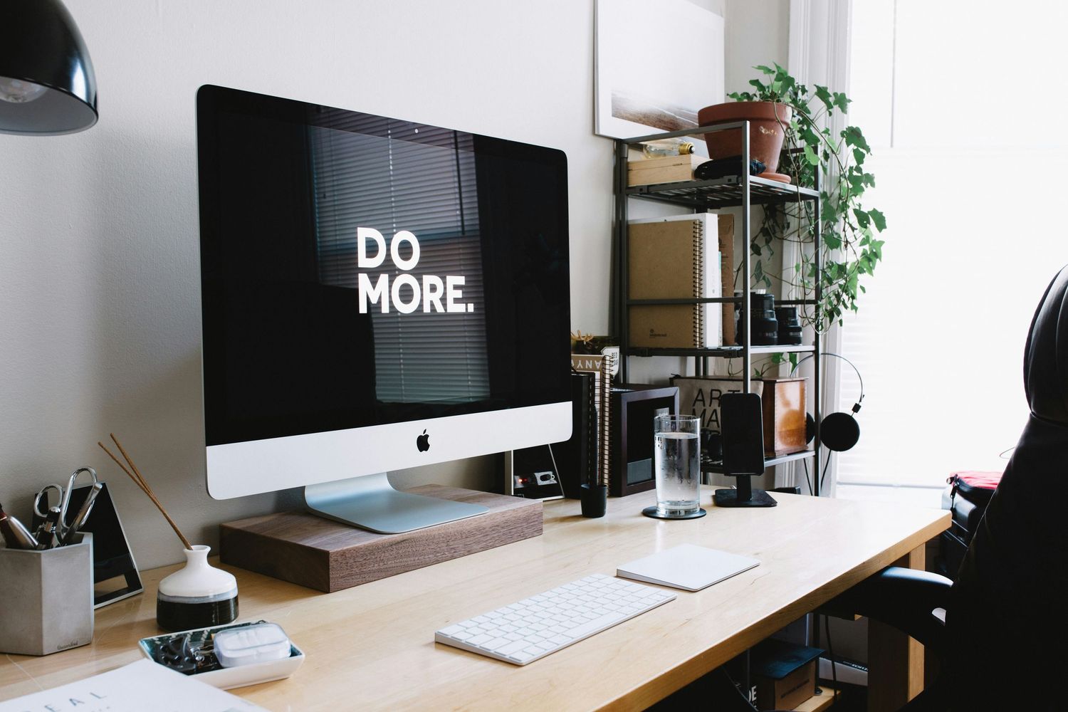 A minimalist home office desk with an iMac displaying 'Do More,' keyboard, plants, and headphones on a shelf.