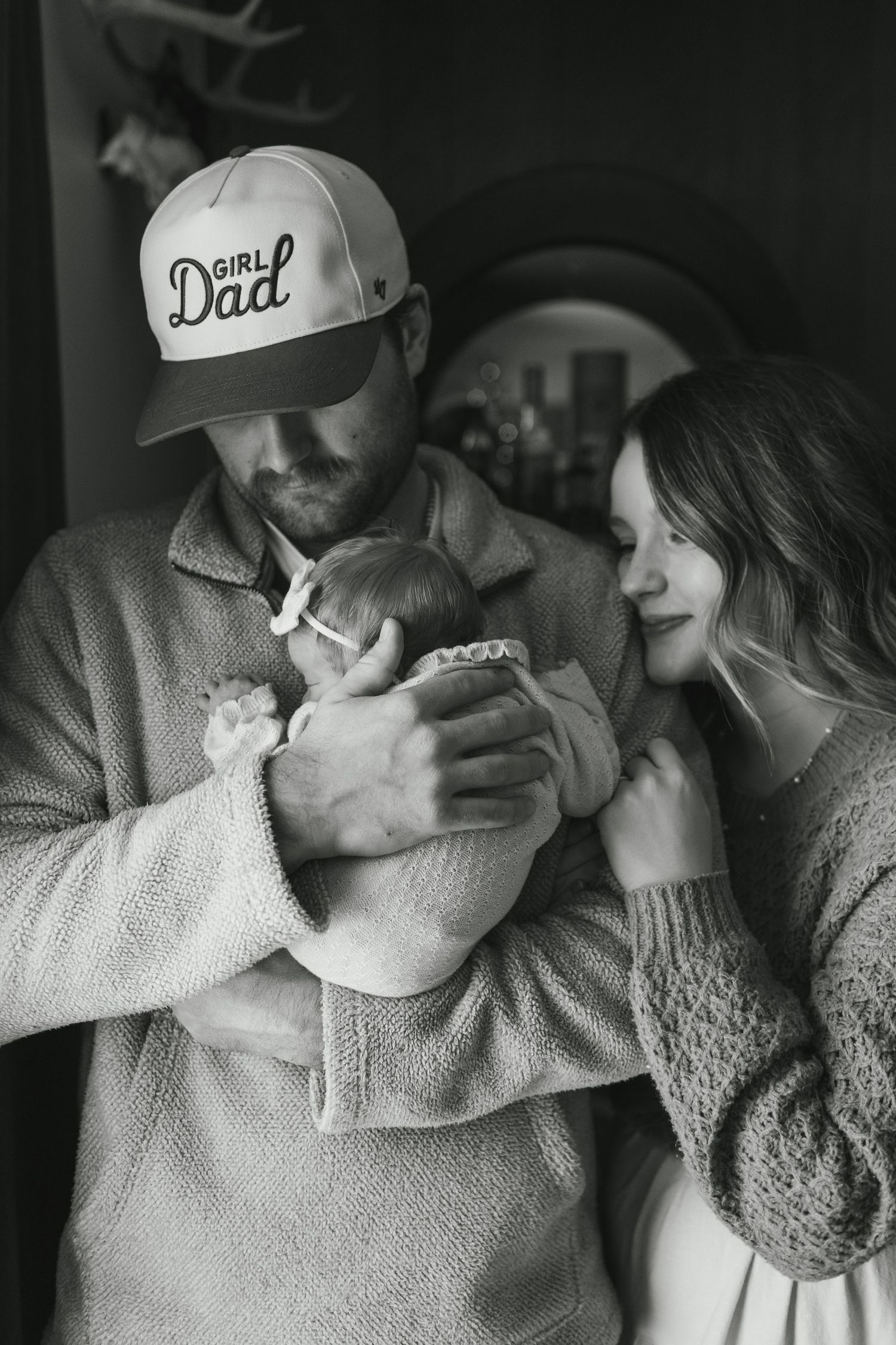 Black and white photo of a couple tenderly holding their newborn baby, father wearing a Girl Dad cap.